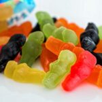 Assorted colourful gummy sweets shaped like small figures are scattered on a white surface, with part of an opened sweet bag in the background. | Andrew T. Austin | Metaphors of Movement