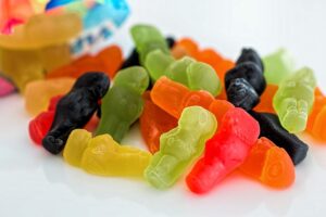 Assorted colourful gummy sweets shaped like small figures are scattered on a white surface, with part of an opened sweet bag in the background. | Andrew T. Austin | Metaphors of Movement