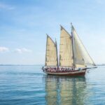 A large sailing boat with three sails carries a group of people across calm, blue water on a clear, sunny day. | Andrew T. Austin | Metaphors of Movement