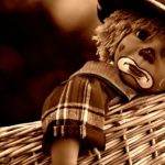 Sepia-toned photo of a sad clown doll with a dummy, curly hair, and checked outfit, resting against a woven basket—a whimsical reminder that even staff development and training can benefit from moments of reflection. | Andrew T. Austin | Metaphors of Movement