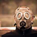 A person wearing a gas mask and dark jacket stands outdoors, embodying Models of Suffering, in front of a blurred background with a wooden fence and red-and-white tape. | Andrew T. Austin | Metaphors of Movement