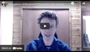 A person with curly hair sits in front of a wooden wall, speaking during a YouTube video titled MoM Discussion: Metaphors of Movement with Andrew T. Austin. | Andrew T. Austin | Metaphors of Movement