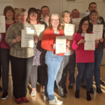 A group of people stand indoors in Langho, holding certificates and smiling at the camera, celebrating their training success. | Andrew T. Austin | Metaphors of Movement