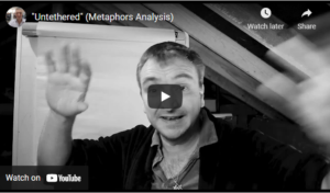 A man with short hair gestures with raised hands in front of a whiteboard in the black and white new video titled Untethered (Metaphor Analysis). | Andrew T. Austin | Metaphors of Movement